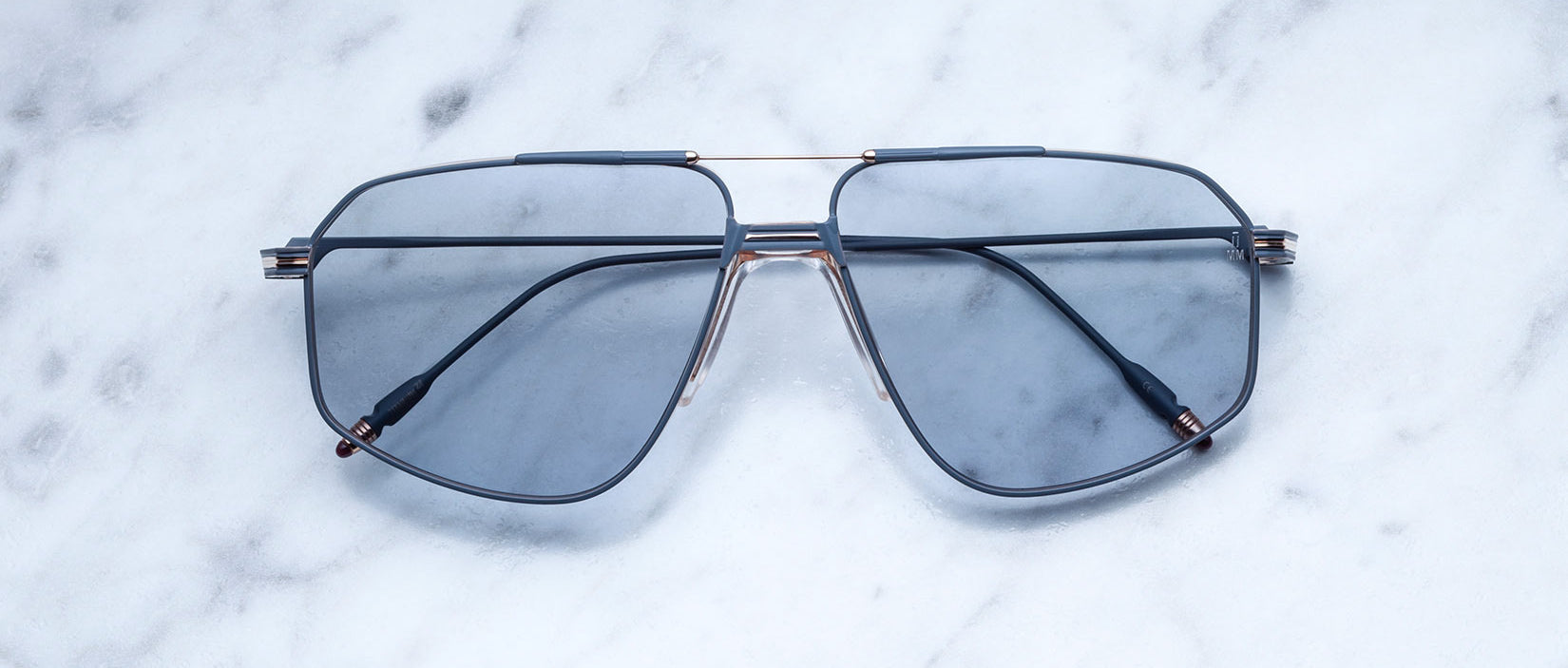 A pair of Jacques Marie Mage Jagger blue-tinted oversized aviator sunglasses with thin beta titanium frames rests on a white marble surface.