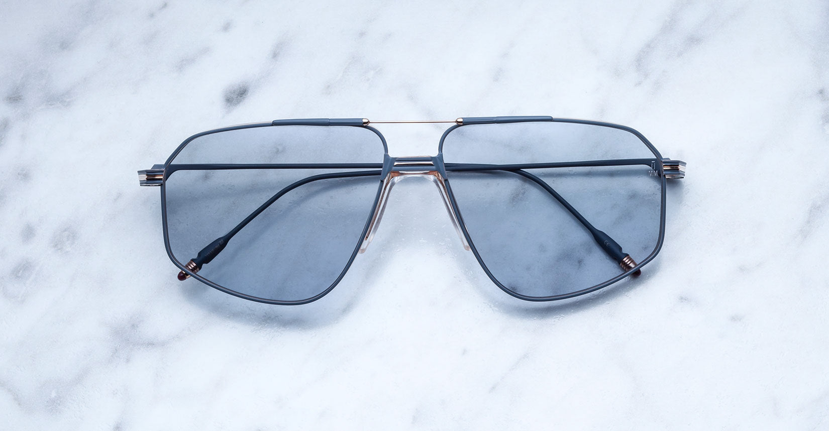 A pair of Jacques Marie Mage Jagger blue-tinted oversized aviator sunglasses with thin beta titanium frames rests on a white marble surface.