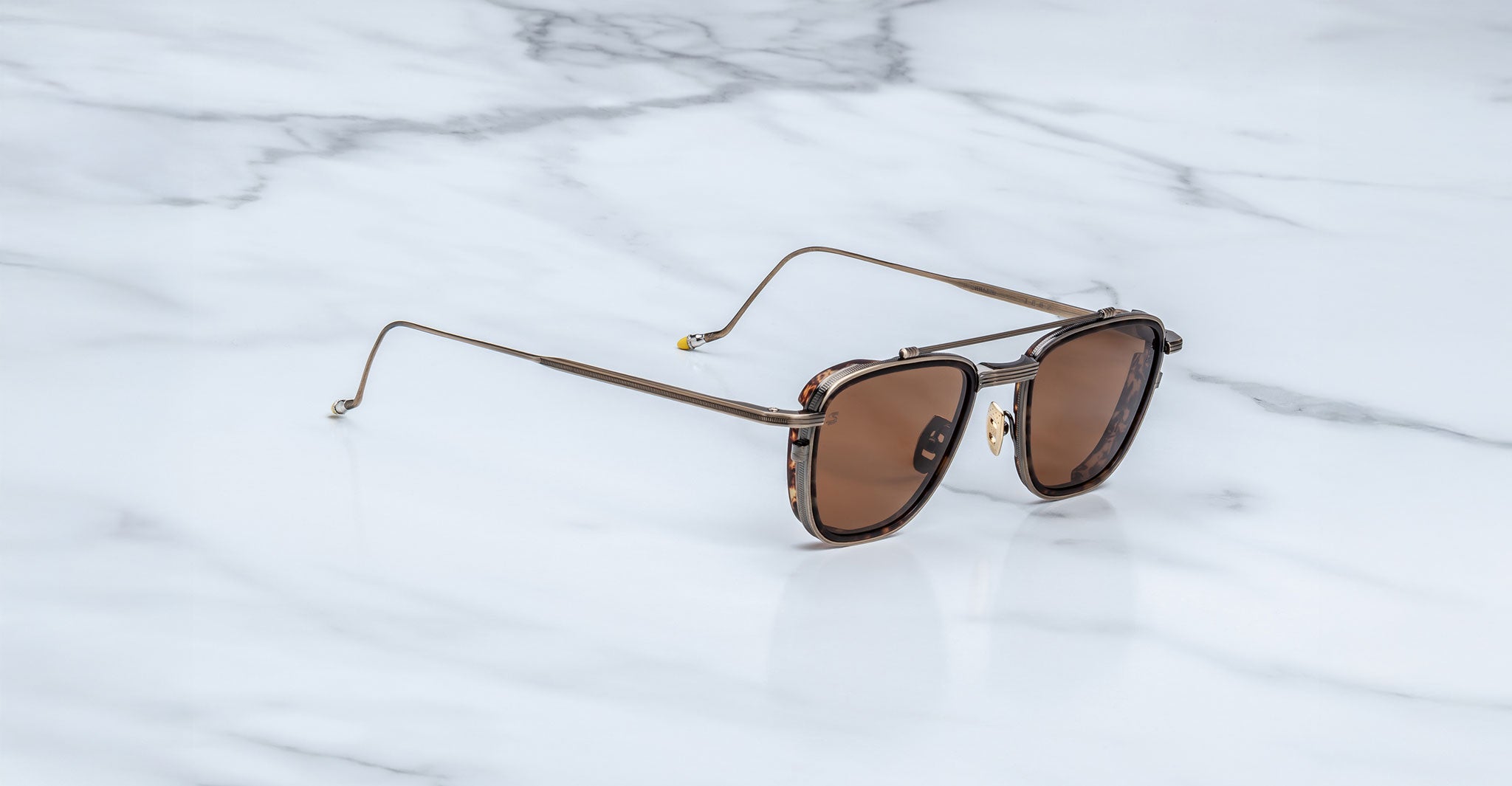 The Jacques Marie Mage 1992 sunglasses feature brown lenses, square frames, and thin gold arms, displayed on a white marble surface.