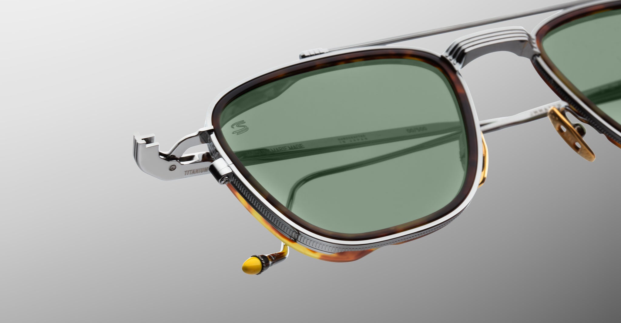 A close-up of Jacques Marie Mage 1992 sunglasses featuring green lenses, metal and tortoiseshell frames, and intricate hinge detailing set against a light gray background.