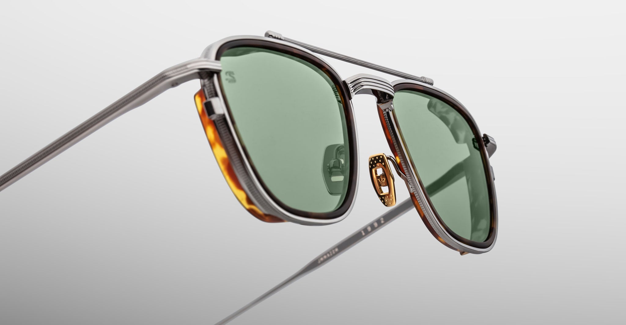 A close-up of Jacques Marie Mage 1992 sunglasses featuring green lenses, silver metal frames, and brown tortoiseshell accents on the rims, set against a plain light gray background.