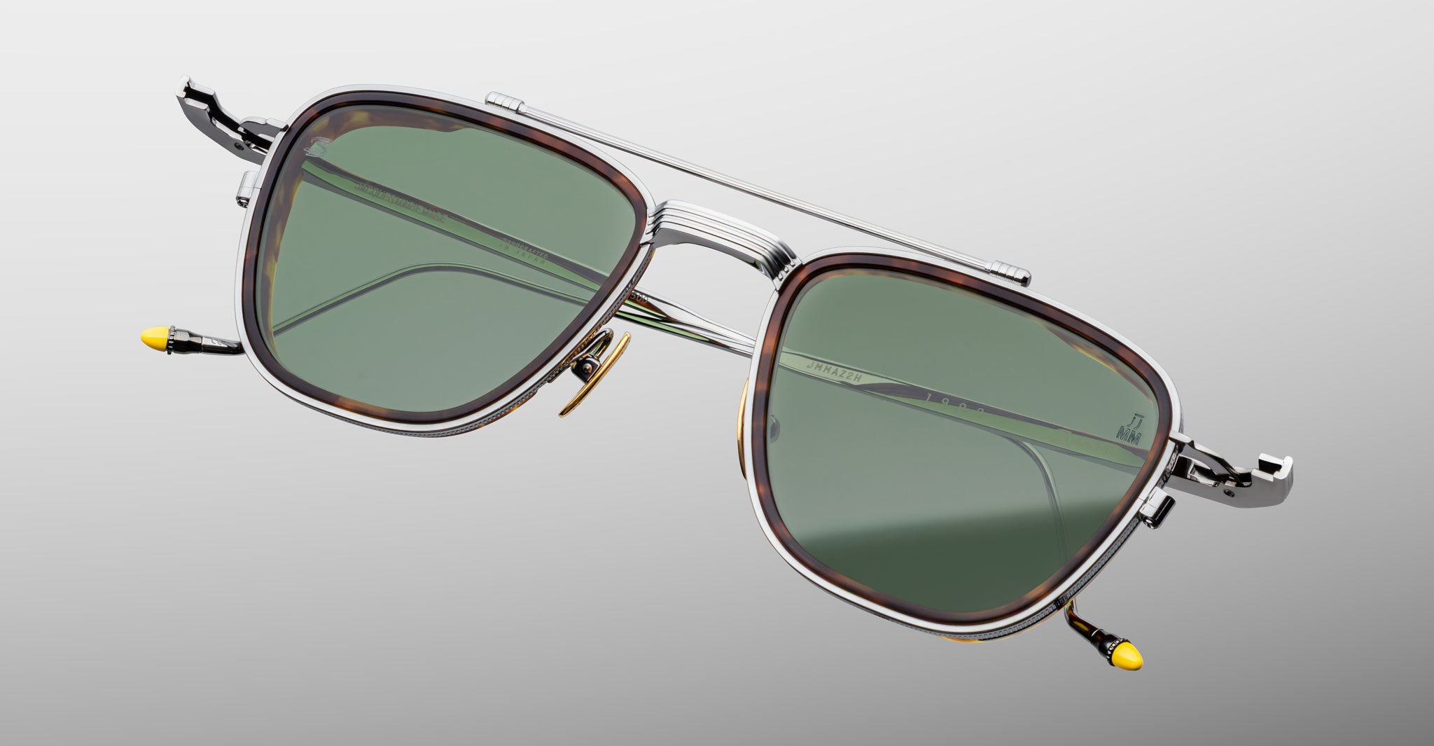 The Jacques Marie Mage 1992 sunglasses feature green-tinted lenses, silver metal frames, and yellow-accented temple tips, shown against a light gray background.