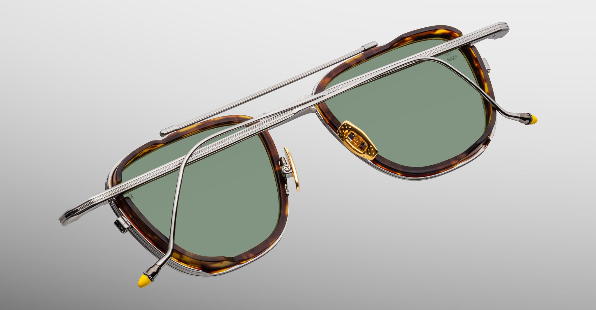 The Jacques Marie Mage 1992 sunglasses feature tortoiseshell frames, green lenses, and unique silver side arms crossed in an X-shape, displayed against a light grey background to showcase their distinctive design.