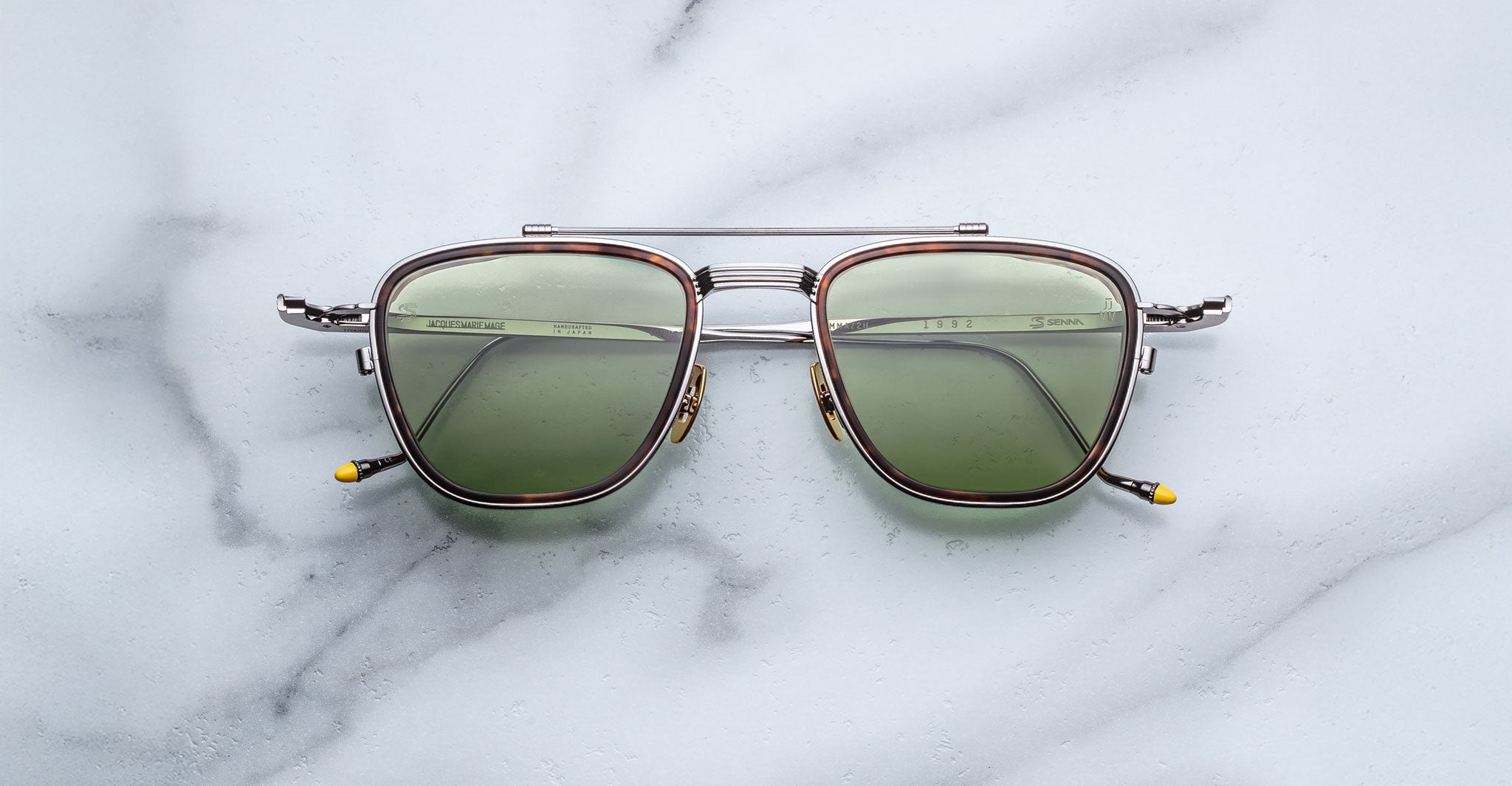 A pair of Jacques Marie Mage 1992 sunglasses with metal square frames, green lenses, thin silver rims, and yellow-tipped arms rests on a light gray marble-like surface.