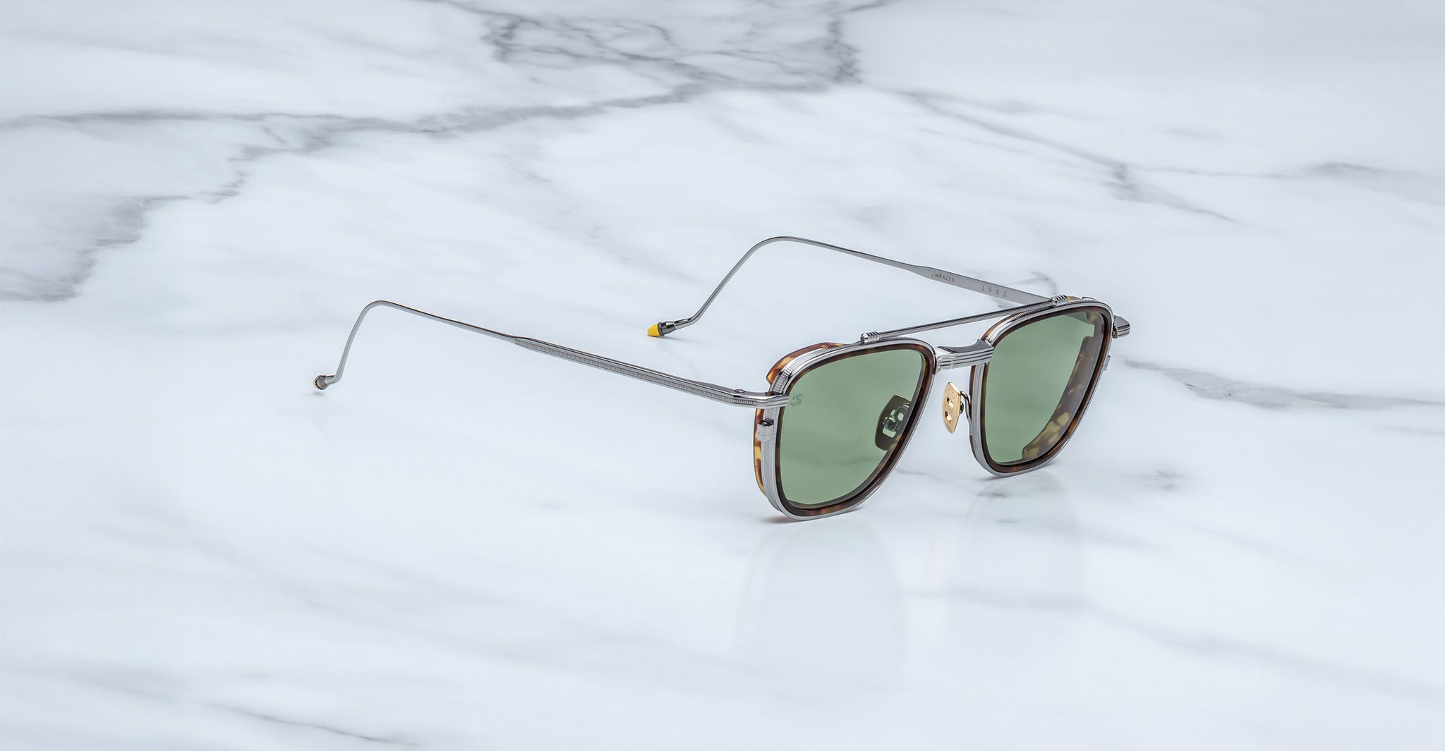 The Jacques Marie Mage 1992 sunglasses, featuring green lenses, thin silver metal arms, and a tortoiseshell frame, rest on a white marble surface with grey veining.