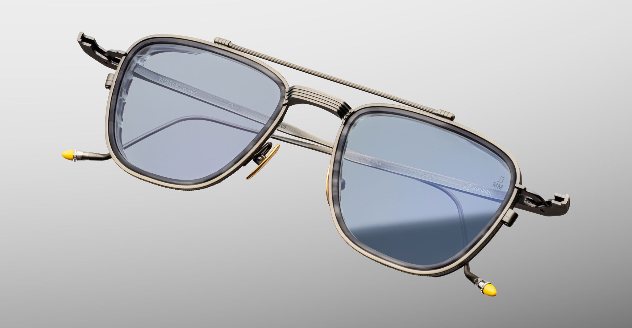 The Jacques Marie Mage 1992 sunglasses feature blue-tinted rectangular lenses, thin metal frames, and yellow temple tip accents, displayed against a light gray background.
