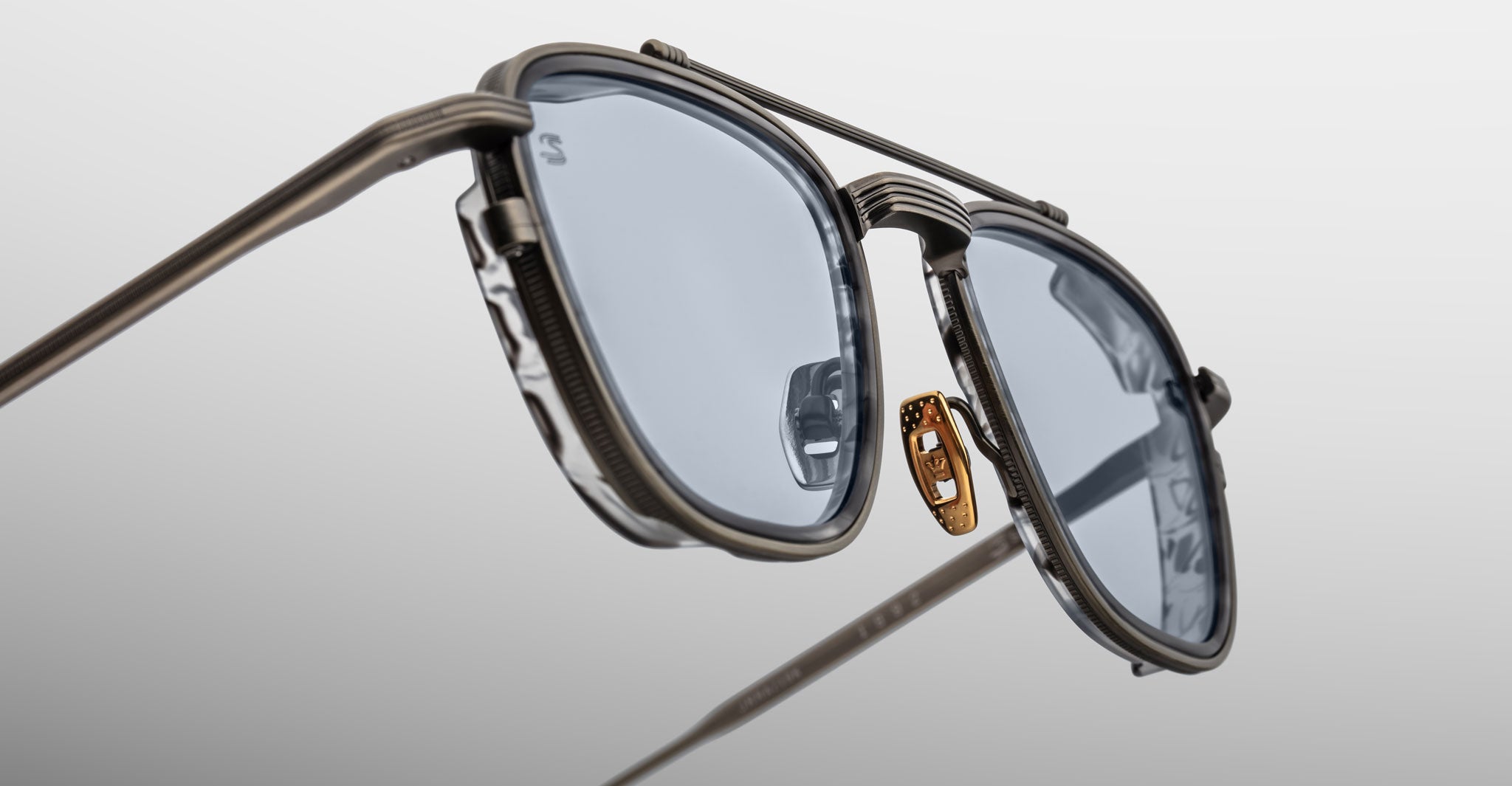 Close-up of Jacques Marie Mage 1992 sunglasses featuring stylish metal frames, blue-tinted lenses, and a gold-toned nose pad, set against a plain gradient gray background.