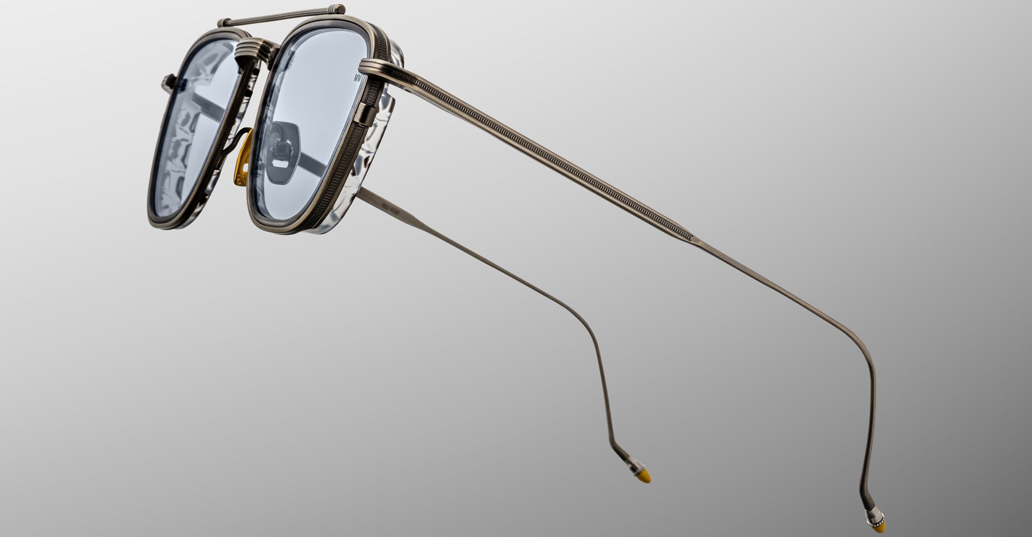 The Jacques Marie Mage 1992 features stylish metal frames with double bridges, thin elongated arms, clear lenses, and yellow-tipped temple ends, all set against a plain gradient background.