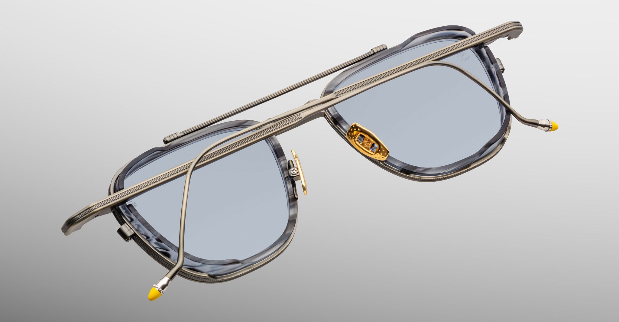 The Jacques Marie Mage 1992 sunglasses feature blue-tinted square lenses, a metallic frame with yellow nose pads and temple tips, and a unique double-bridge design, showcased against a gradient gray background.