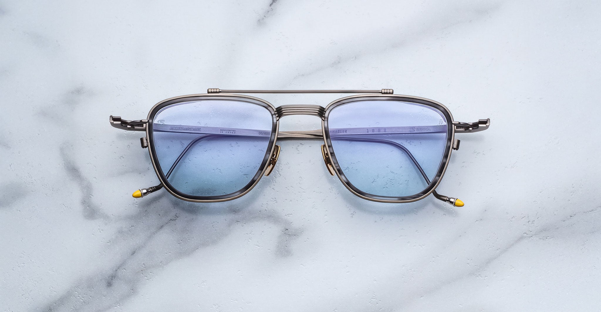 The Jacques Marie Mage 1992 sunglasses, featuring square metal frames with fine details, blue-tinted lenses, and yellow-tipped arms, are displayed on a white marble surface.