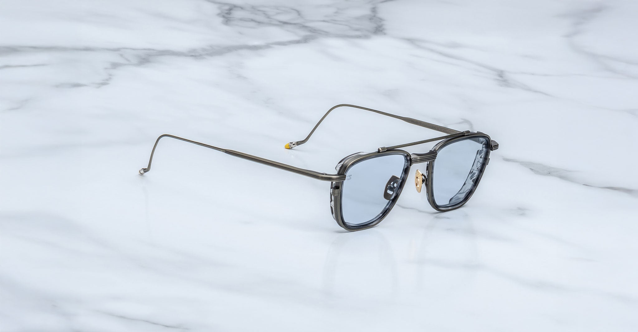 The Jacques Marie Mage 1992 sunglasses feature silver rims, blue-tinted lenses, and thin metal arms, displayed on a white marble surface.
