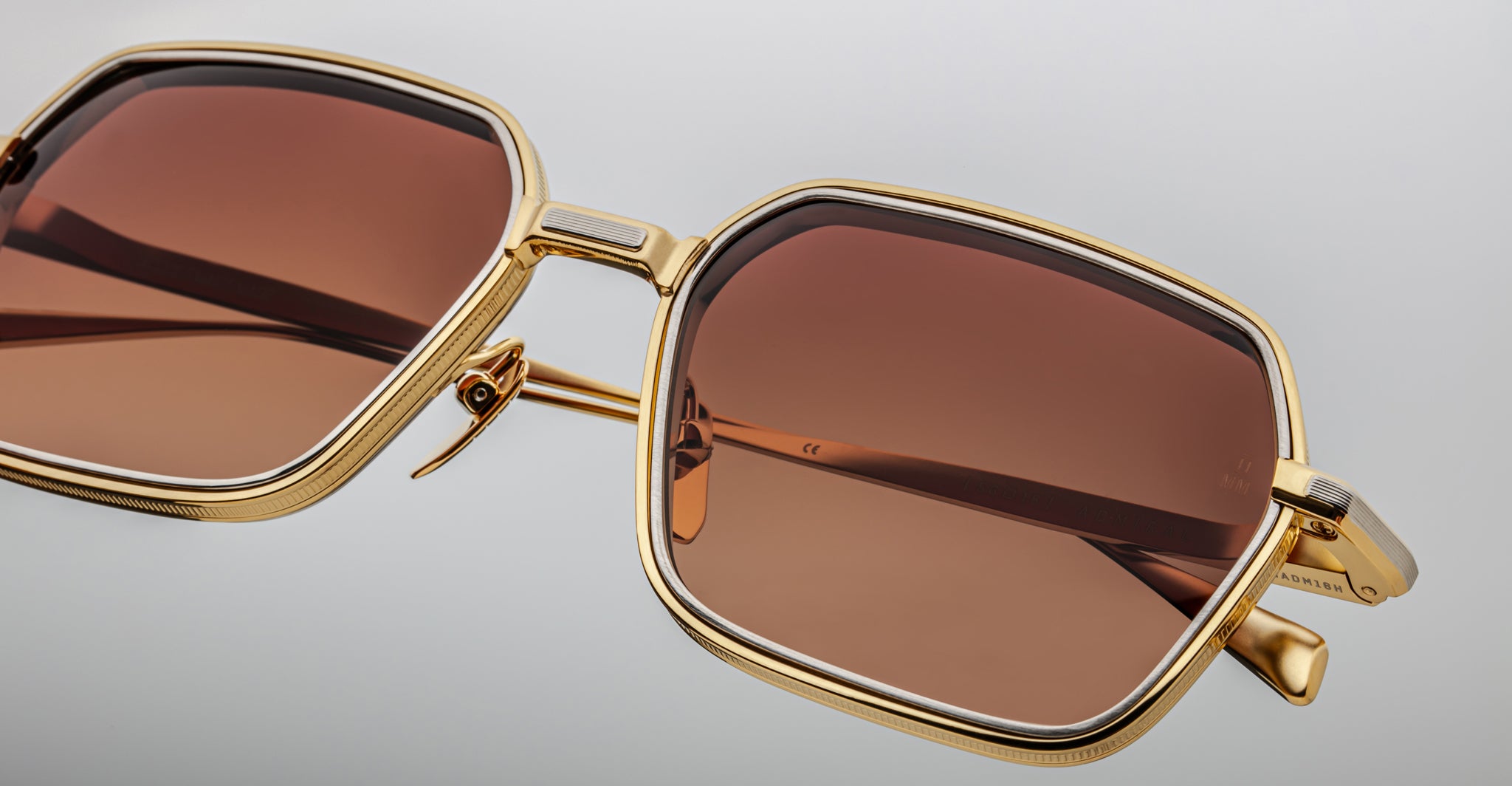 Close-up of Jacques Marie Mage Admiral sunglasses featuring stylish brown-tinted square lenses and gold metal frames, displayed against a light gray background.