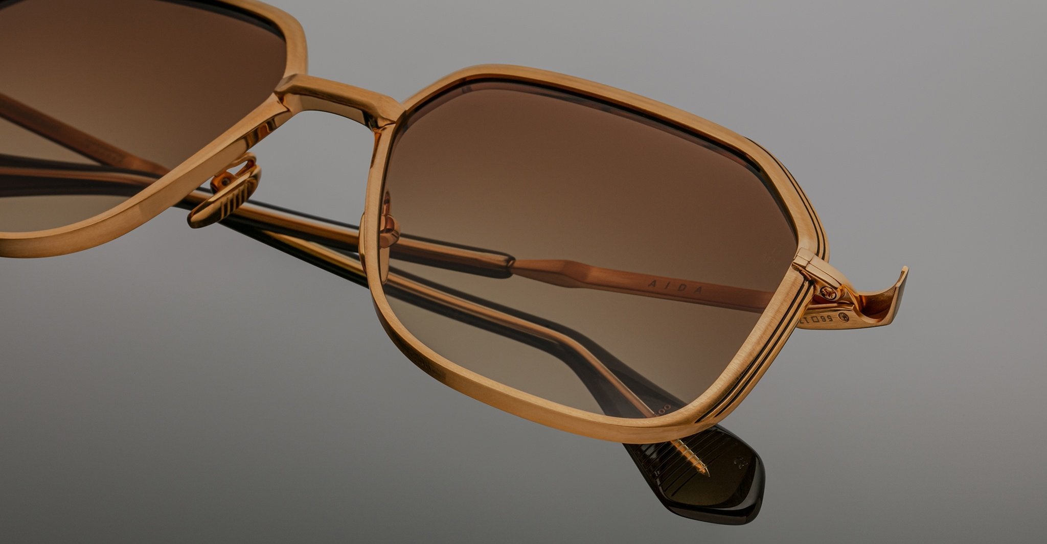 Angled close-up of Jacques Marie Mage Aida sunglasses in gold-tone metal, highlighting the geometric lenses with brown gradient tint, sculpted bridge, engraved temple, and black acetate temple tip.