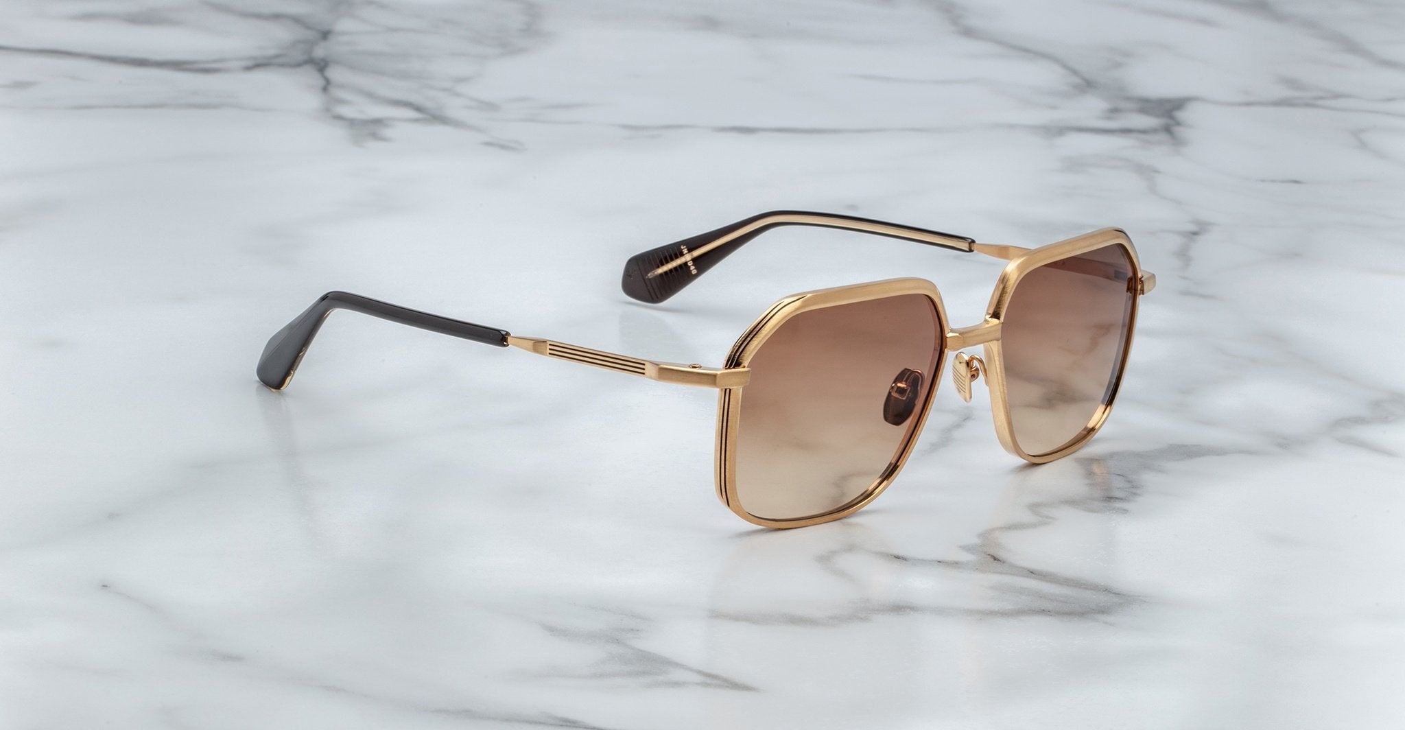 Angled full view of Jacques Marie Mage Aida sunglasses in gold-tone metal, featuring geometric square lenses with brown gradient tint, sculpted bridge, engraved temples, and black acetate temple tips on a marble surface.