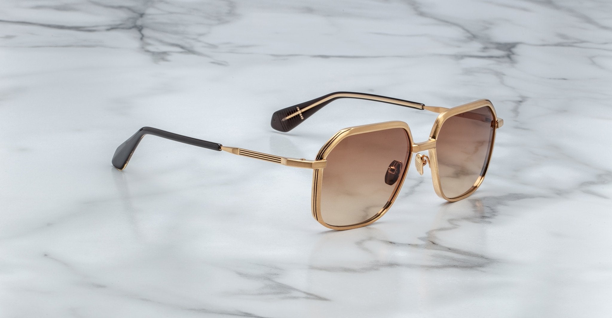 Angled full view of Jacques Marie Mage Aida sunglasses in gold-tone metal, featuring geometric square lenses with brown gradient tint, sculpted bridge, engraved temples, and black acetate temple tips on a marble surface.