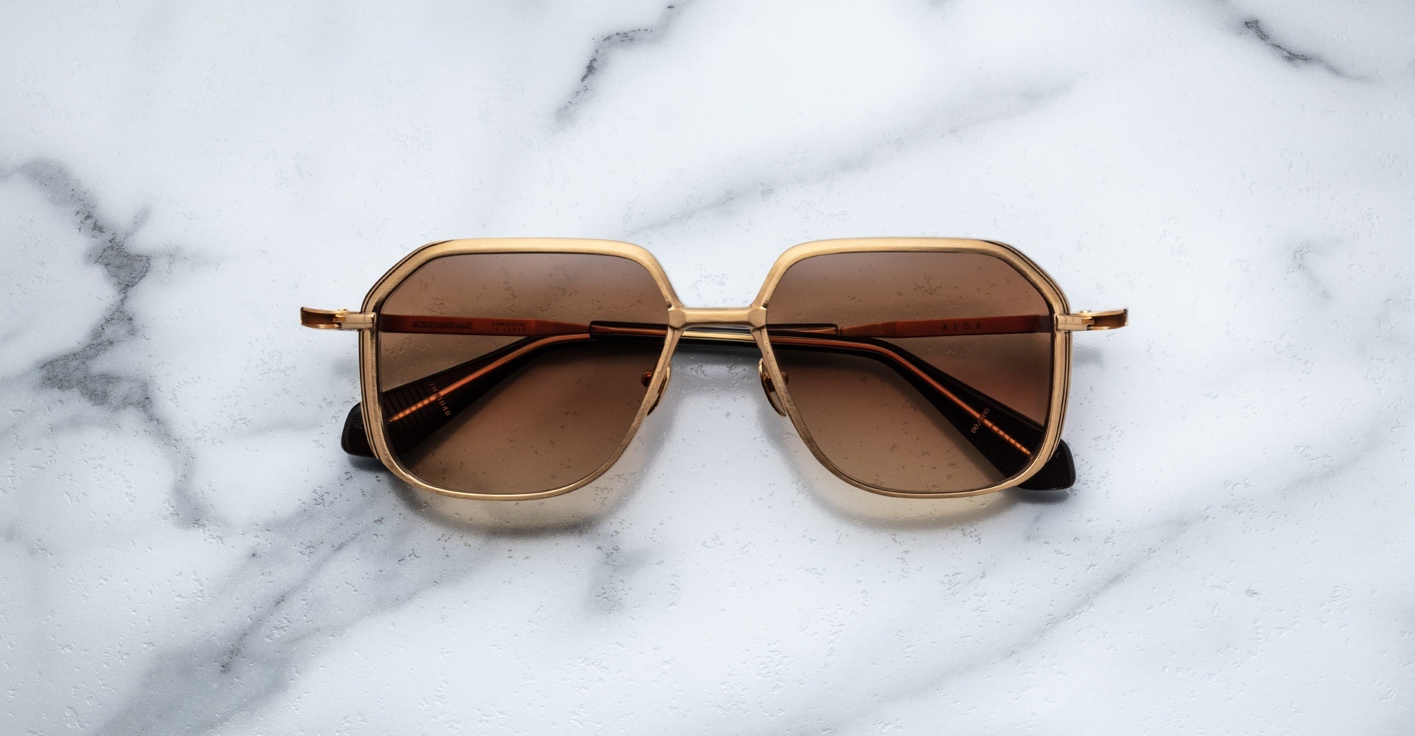 Jacques Marie Mage Aida sunglasses in gold-tone metal with a geometric square aviator shape, double bridge, and brown tinted lenses, front view on a neutral background.