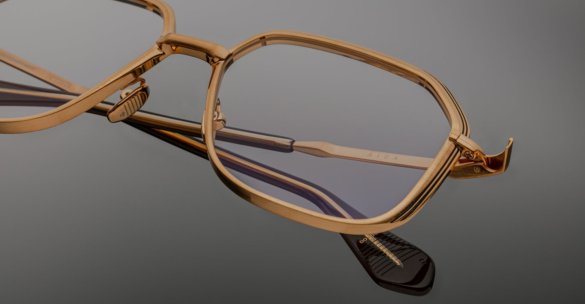Angled close-up of Jacques Marie Mage Aida eyeglasses in gold-tone metal, highlighting the faceted rim, double bridge, engraved temple, and black acetate temple tip.