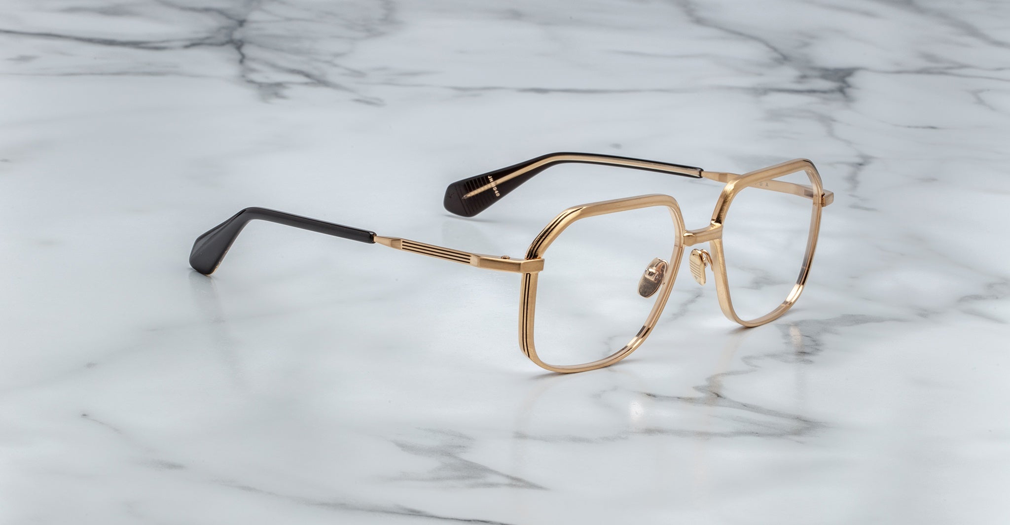 Angled full view of Jacques Marie Mage Aida eyeglasses in gold-tone metal, featuring geometric square lenses, a sculpted bridge, engraved temples, and black acetate temple tips on a marble surface.