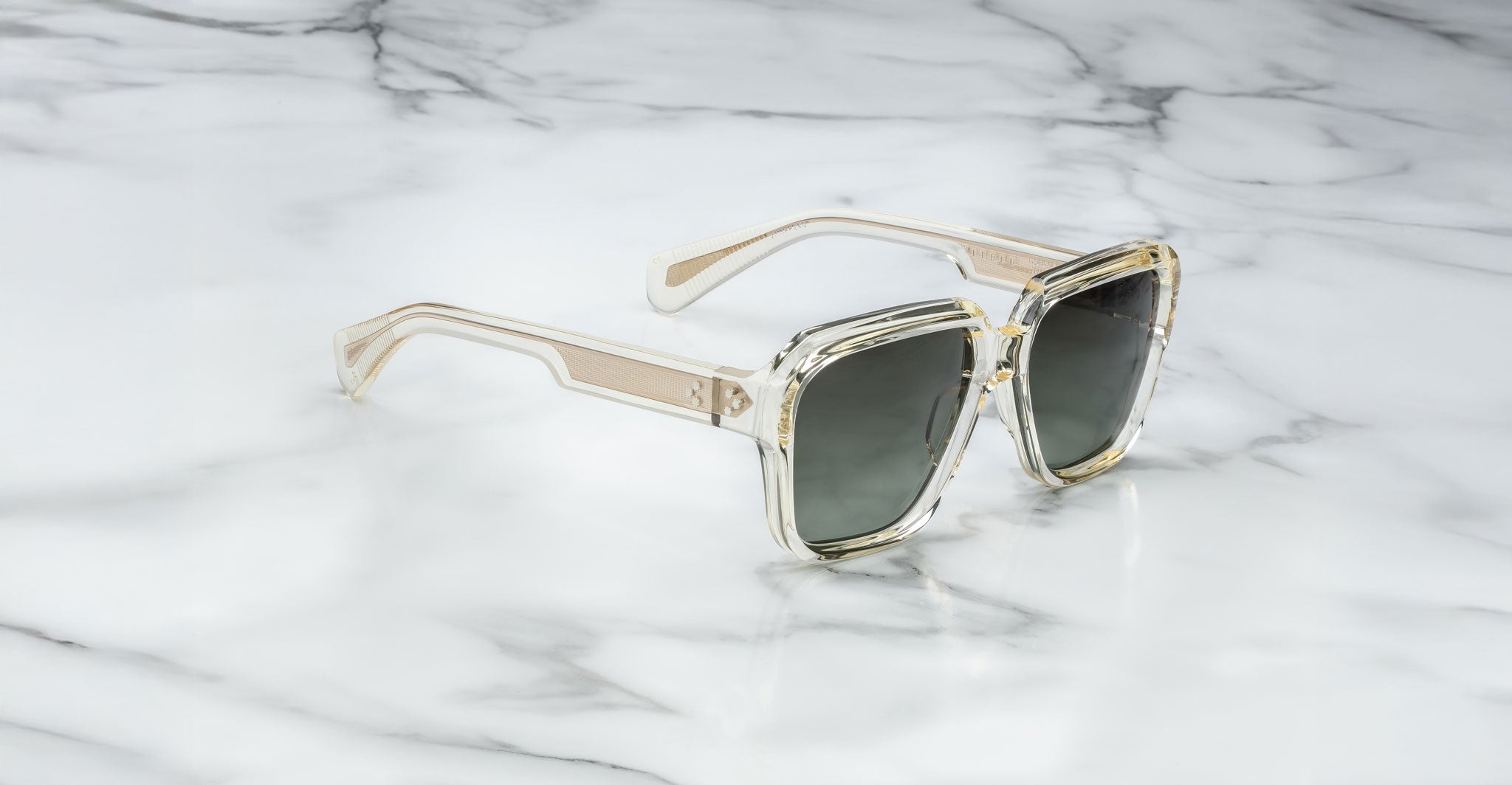 A pair of Jacques Marie Mage Alfred square sunglasses with clear, oversized frames and dark lenses rests on a white marble surface.