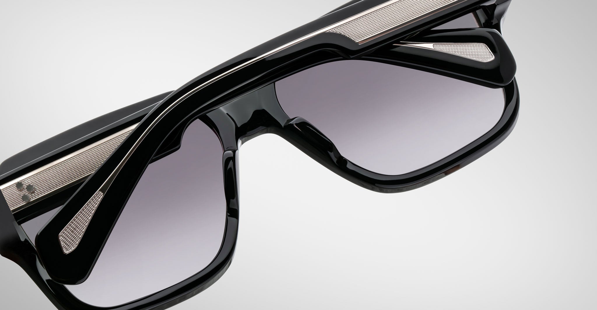 A close-up of Jacques Marie Mage Alfred sunglasses highlights their thick black frames, silver metal accents, and gradient gray lenses against a plain light background.
