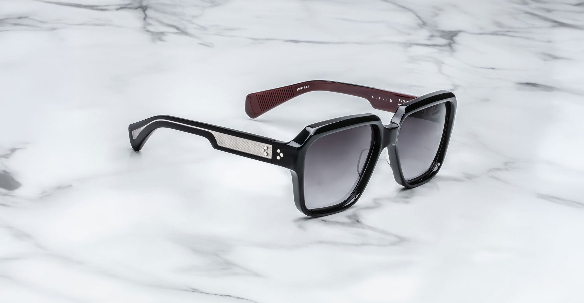 The Jacques Marie Mage Alfred sunglasses feature a black rectangular frame with silver detailing on the temples, maroon textured inner arms, and dark, slightly reflective lenses, displayed on a white marble surface.