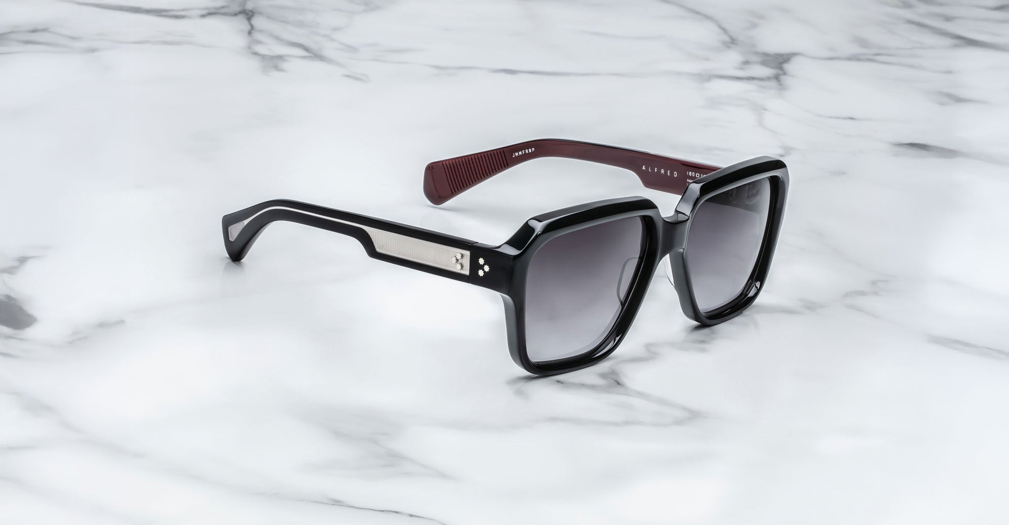 The Jacques Marie Mage Alfred sunglasses feature a black rectangular frame with silver detailing on the temples, maroon textured inner arms, and dark, slightly reflective lenses, displayed on a white marble surface.