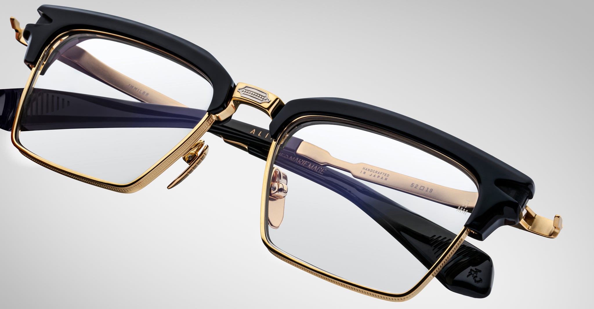Close-up of Jacques Marie Mage Alin eyeglasses featuring black and gold frames, clear lenses, and detailed hinges. The arms display embossed text and intricate design elements, set against a plain light background.