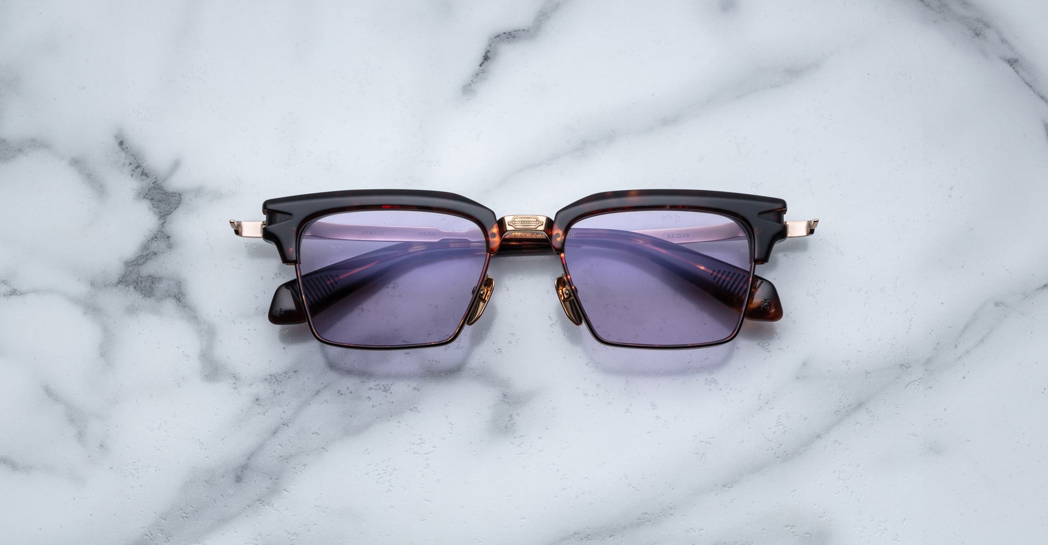 The Jacques Marie Mage Alin sunglasses feature dark purple lenses and a black and brown frame, displayed on a white marble surface.
