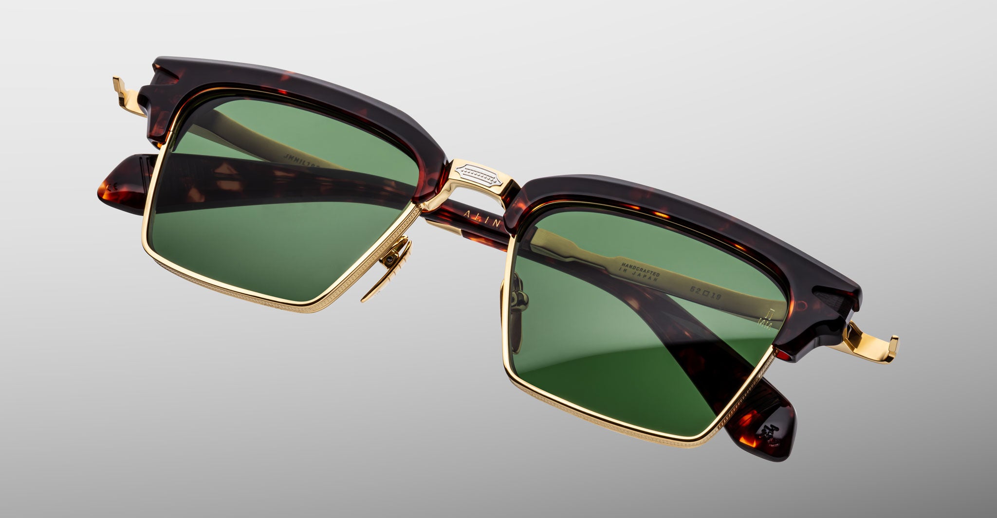 The Jacques Marie Mage Alin sunglasses feature green square lenses, gold metal accents, and a dark tortoiseshell brown frame, displayed against a plain gray background.