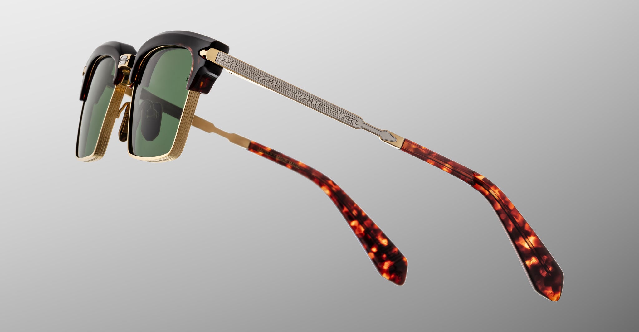 The Jacques Marie Mage Alin sunglasses feature green lenses, gold and black frames, patterned silver arms, and brown tortoiseshell temple tips, displayed against a gray background.