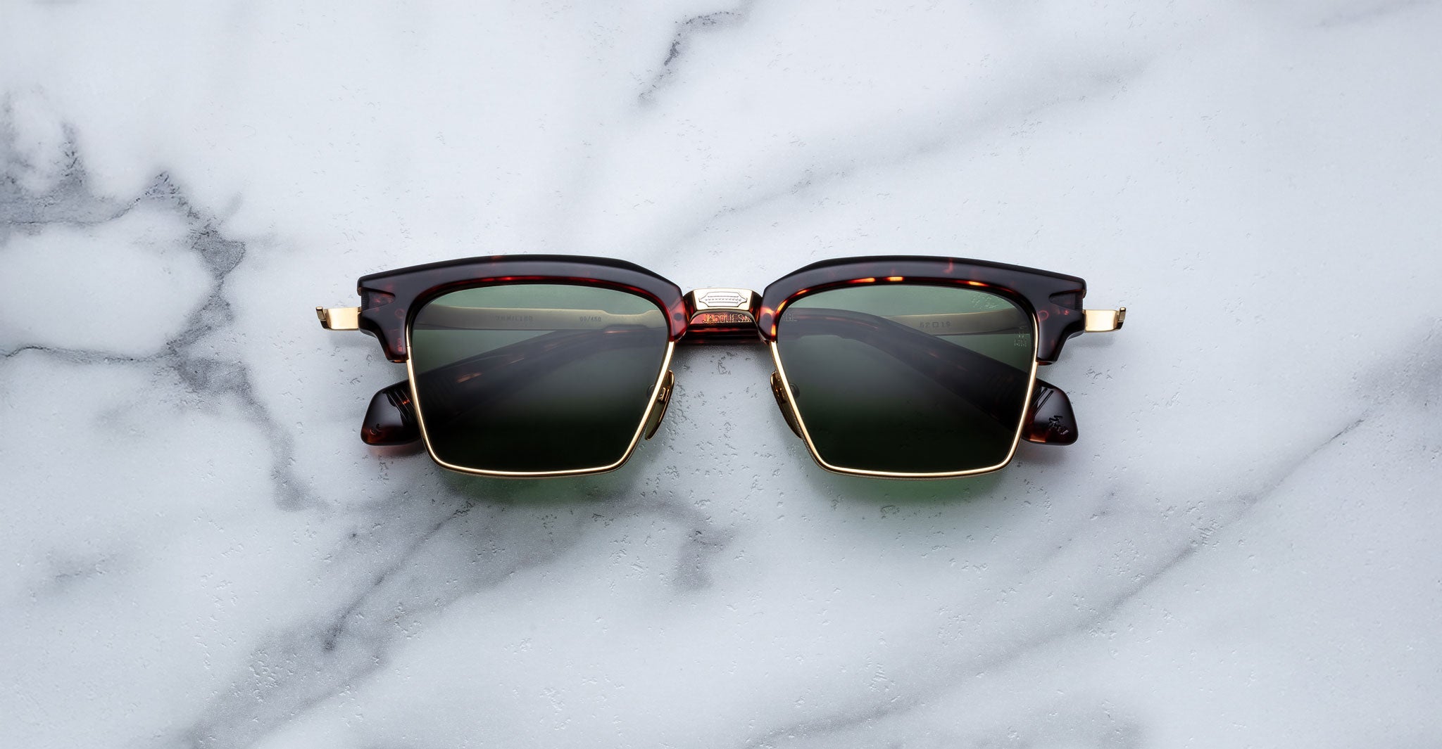 The Jacques Marie Mage Alin sunglasses, featuring tortoiseshell and gold frames with dark green lenses, rest on a white marble surface.