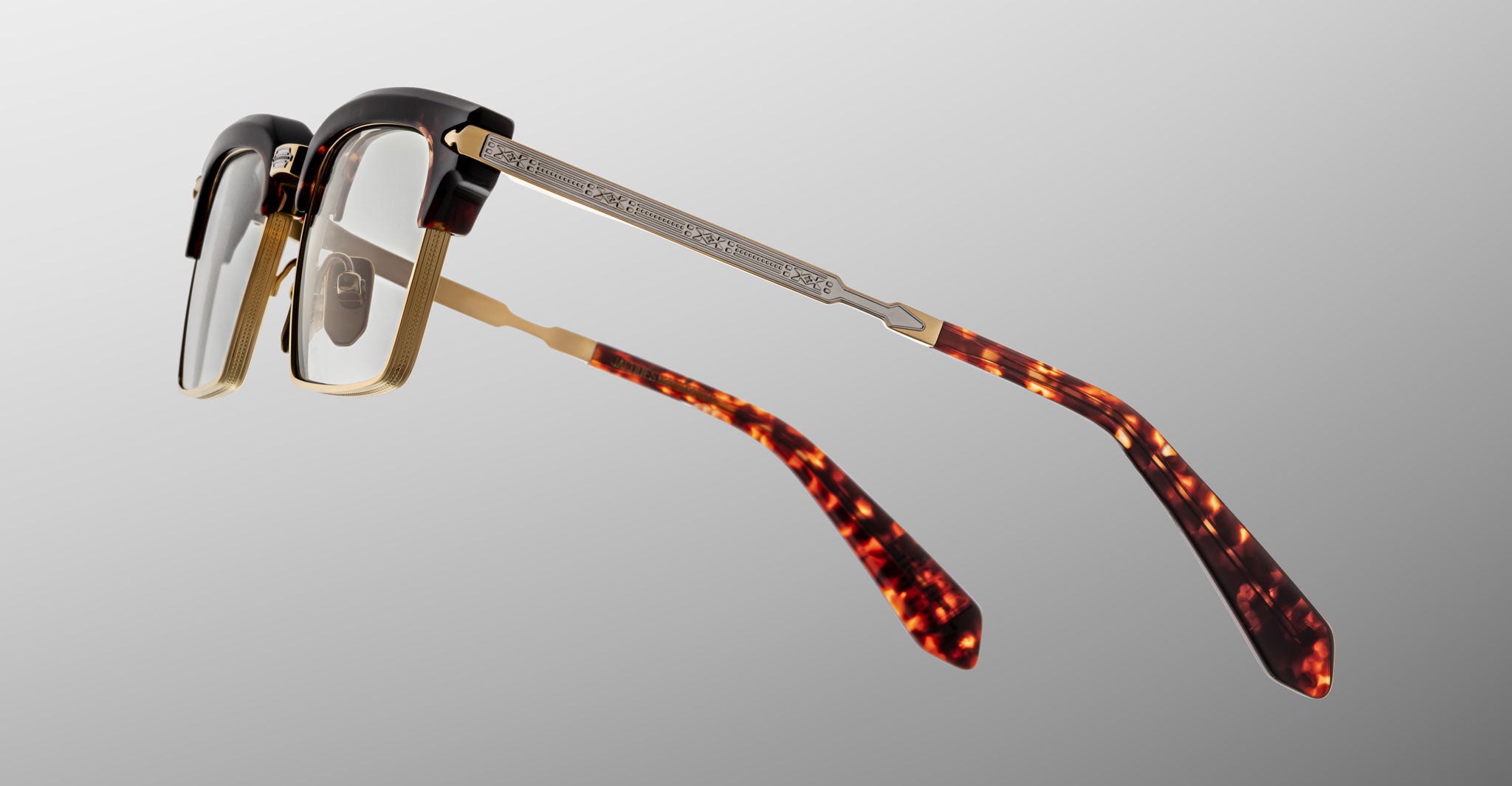 The Jacques Marie Mage Alin eyeglasses feature clear lenses, a black and gold frame, and tortoiseshell-patterned temples, displayed from a rear side angle against a neutral gray background.
