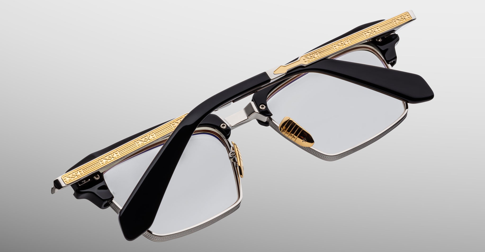 The Jacques Marie Mage Alin eyeglasses feature black arms, gold detailing on the top frame edge, clear lenses, and a bold angular design, displayed against a light gray background.