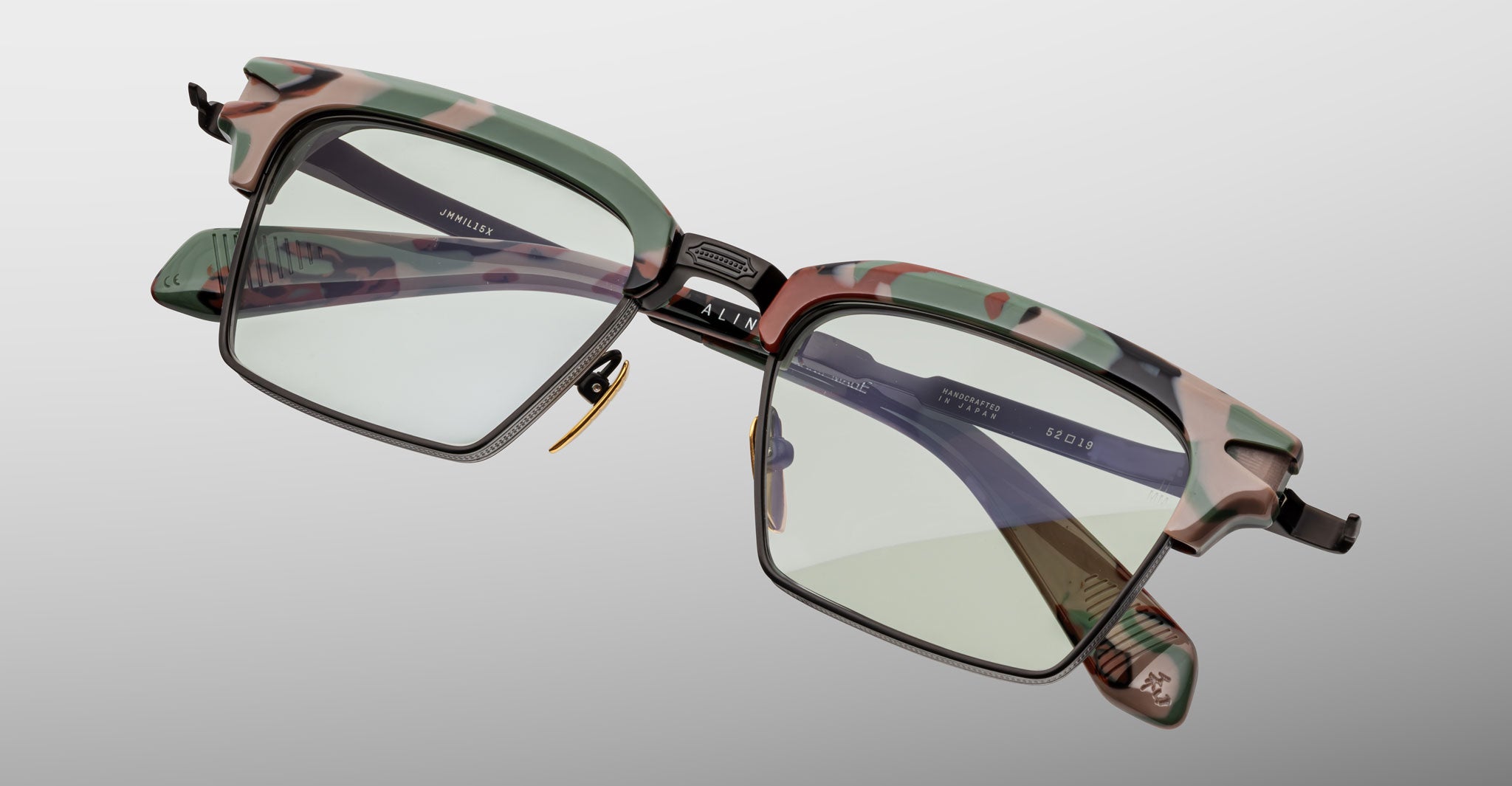 The Jacques Marie Mage Alin eyeglasses feature a green and pink camouflage frame, clear lenses, black lower rims, and metal bridge details, displayed against a light gray background.