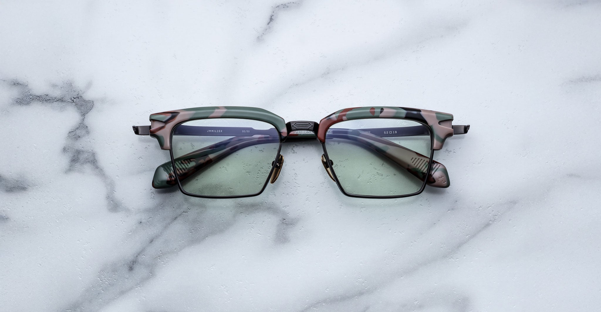 A pair of Jacques Marie Mage Alin square eyeglasses, featuring green and pink camouflage patterns and slightly tinted lenses, is displayed on a white marble surface.