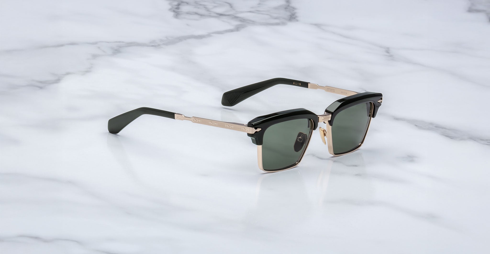 A pair of Jacques Marie Mage Alin black and gold square sunglasses with dark lenses rests on a white marble surface.