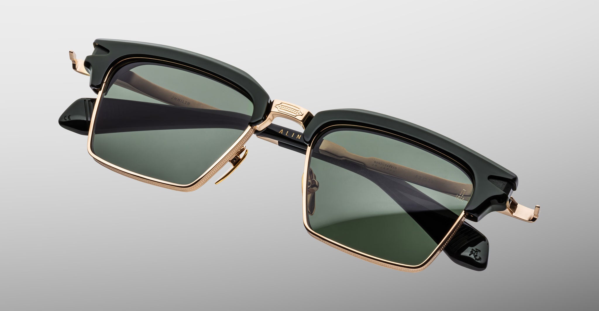 The Jacques Marie Mage Alin features square-shaped sunglasses with dark green lenses, a black upper frame, and gold metal accents, displayed against a light gray gradient background.