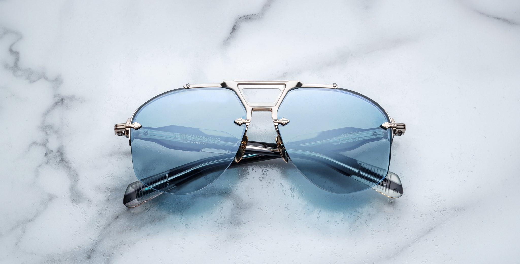 Jacques Marie Mage Alta aviator sunglasses with silver frames and blue-tinted lenses, viewed from above.