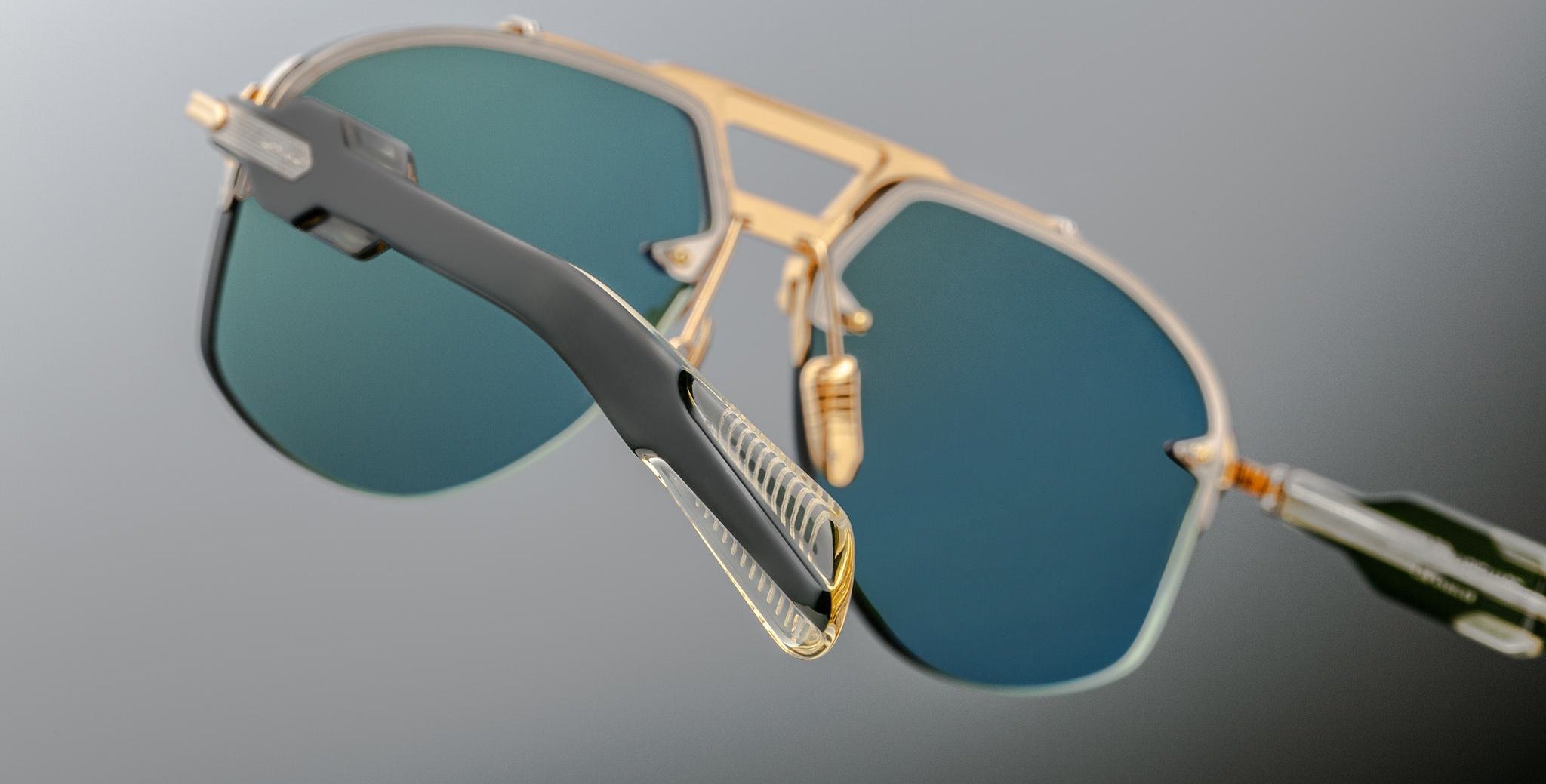 Jacques Marie Mage Alta sunglasses with gold frames, blue-green lenses, and clear arms shown from the front.