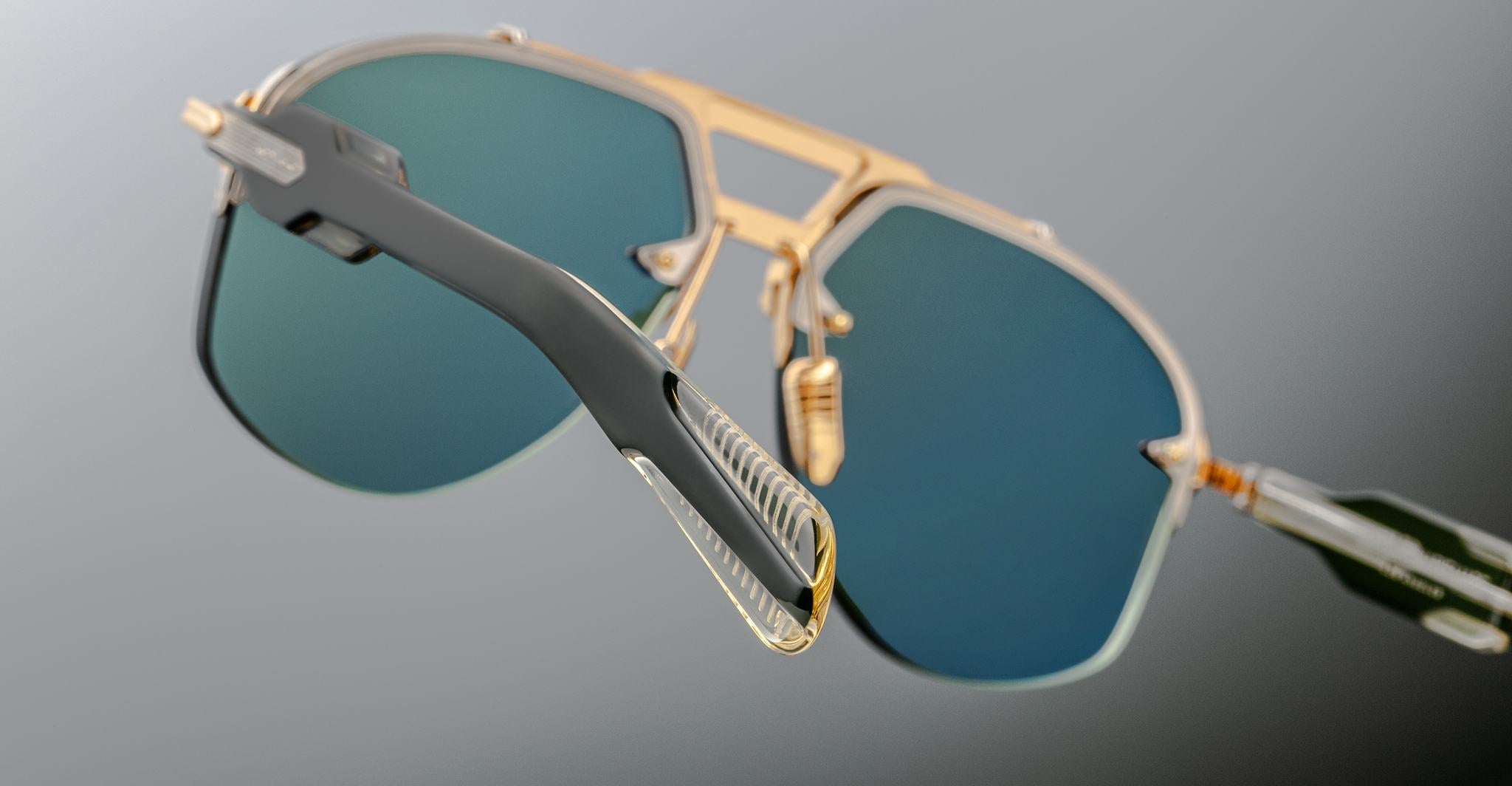 Jacques Marie Mage Alta sunglasses with gold frames, blue-green lenses, and clear arms shown from the front.