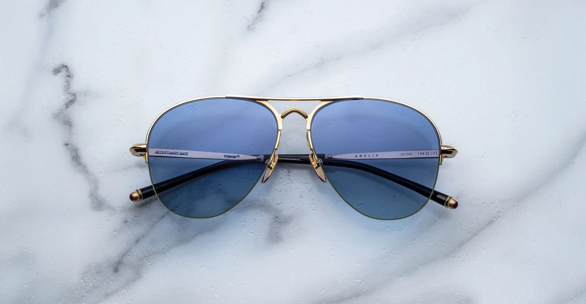 The Jacques Marie Mage Amelia aviator-style sunglasses with blue-tinted lenses and gold metal frames are displayed on a white marble surface with subtle grey veining.