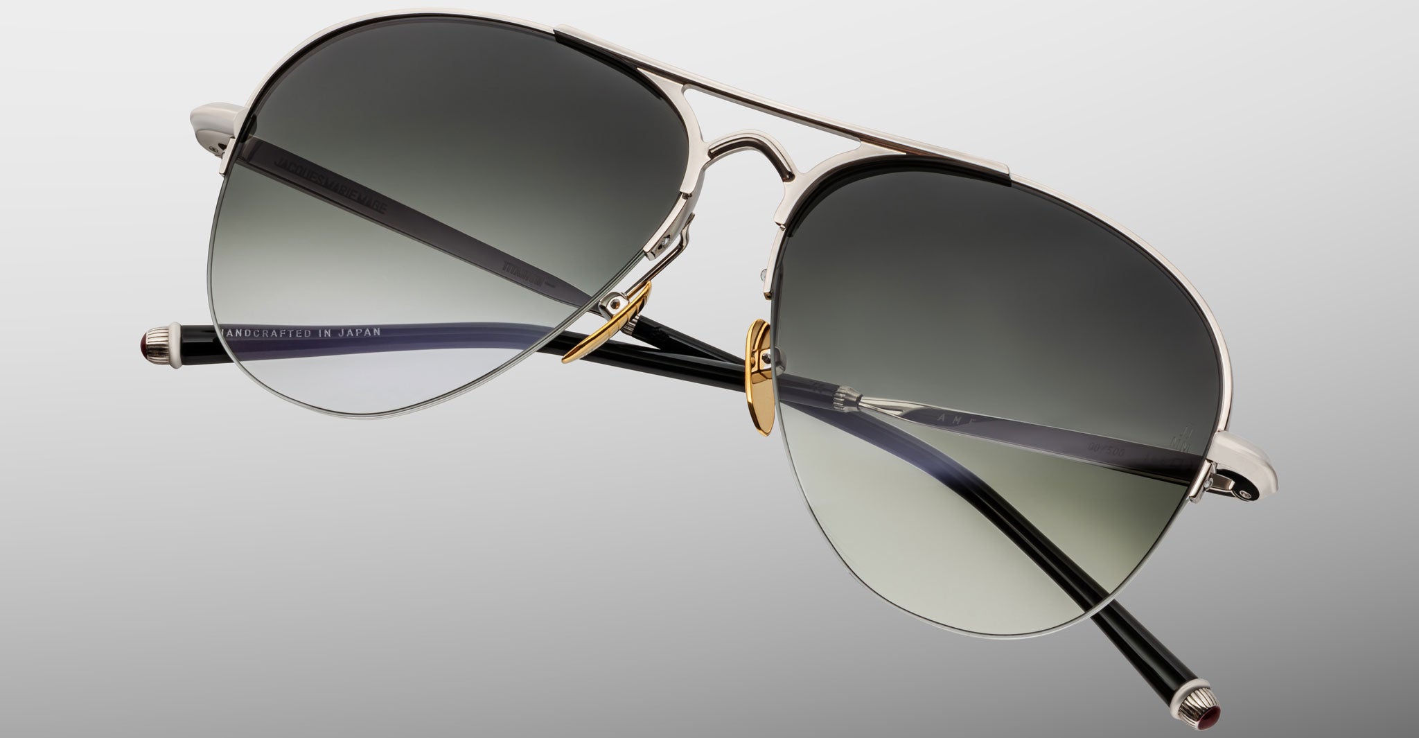 The Jacques Marie Mage Amelia sunglasses feature aviator-style silver metal frames, dark gradient lenses, and black temples, displayed against a light gray background.