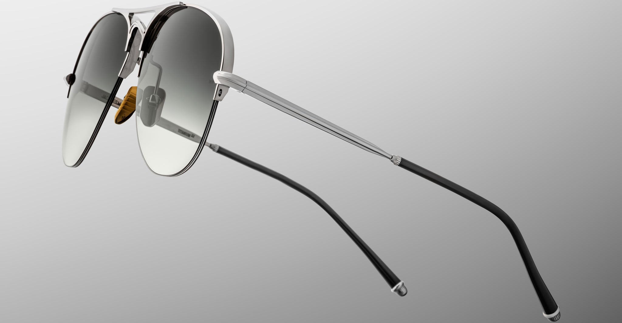 A close-up of Jacques Marie Mage Amelia aviator sunglasses featuring metal frames, gradient gray lenses, and black temple tips, set against a plain gray background.