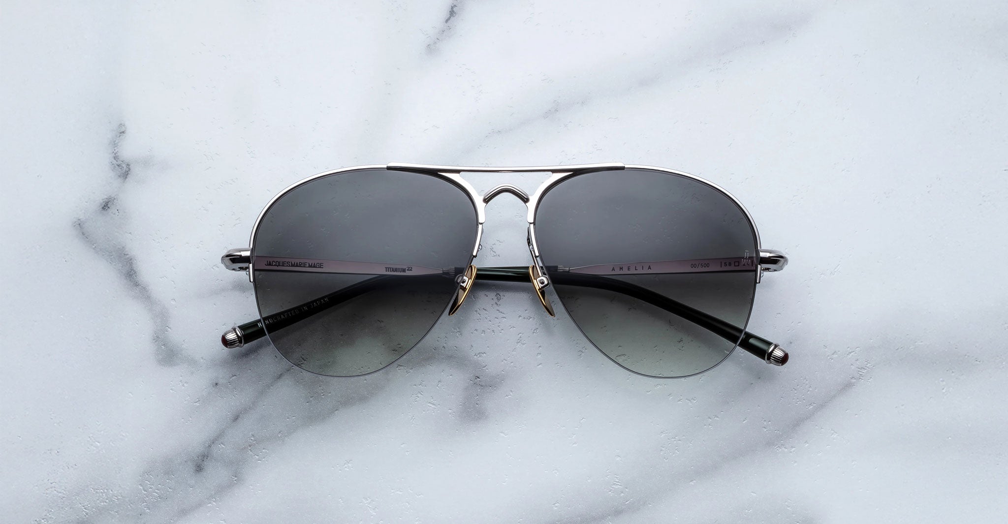 The Jacques Marie Mage Amelia aviator-style sunglasses with dark lenses and silver frames are displayed on a white marble surface, viewed from above.