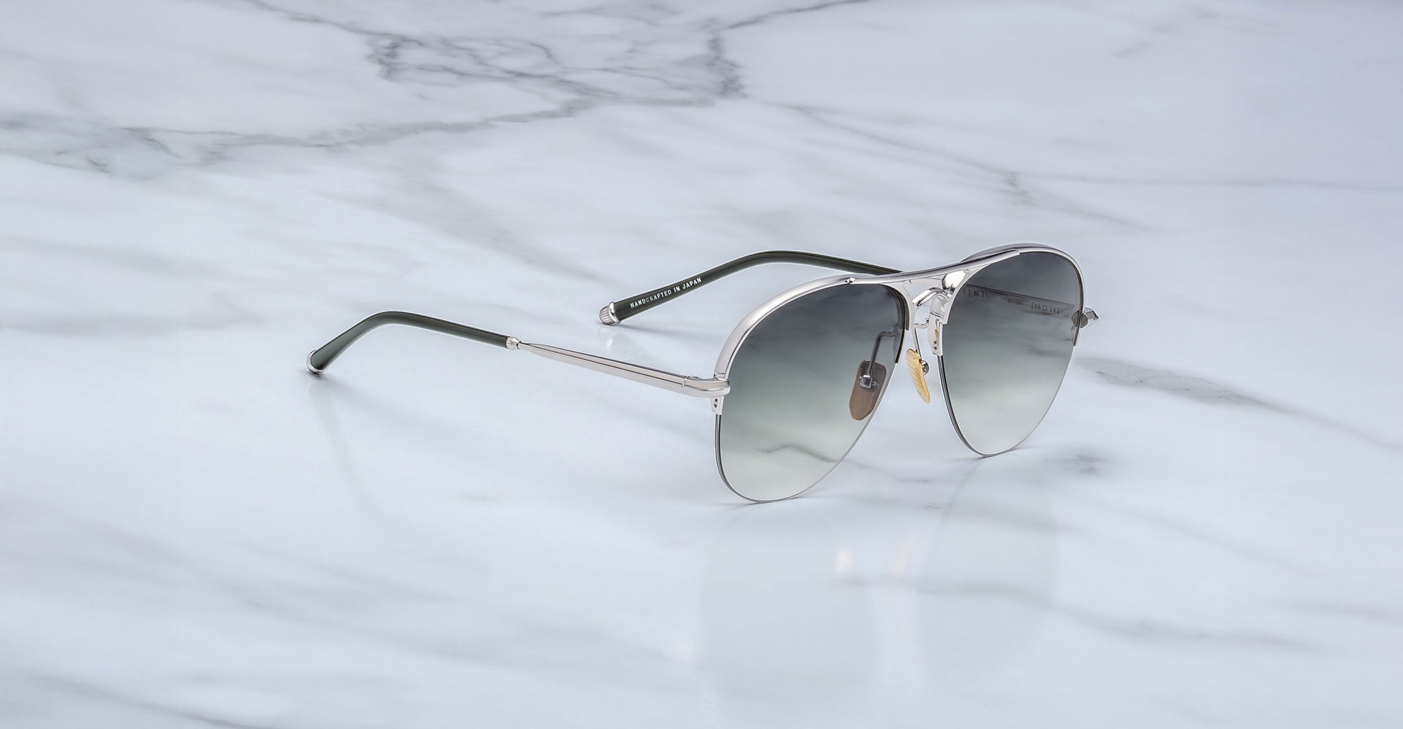 The Jacques Marie Mage Amelia aviator-style sunglasses with silver metal frames and gray gradient lenses rest on a white marble surface.