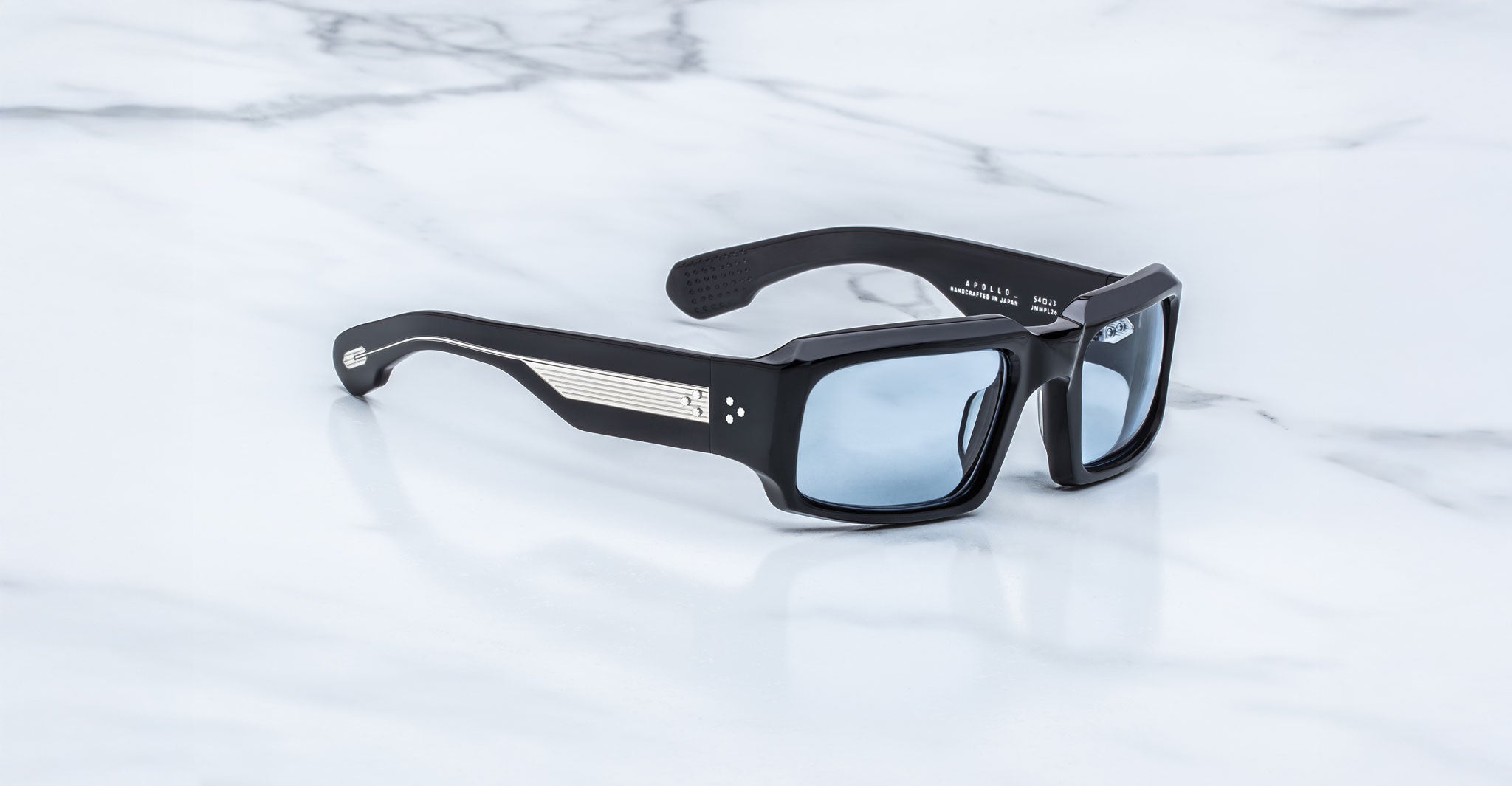 Jacques Marie Mage Apollo in Shadow shown in angled view, featuring a bold rectangular black acetate frame, thick sculpted temples with exposed engraved metal core, triple dot rivets, signature arrowhead tips, and blue-tinted lenses.