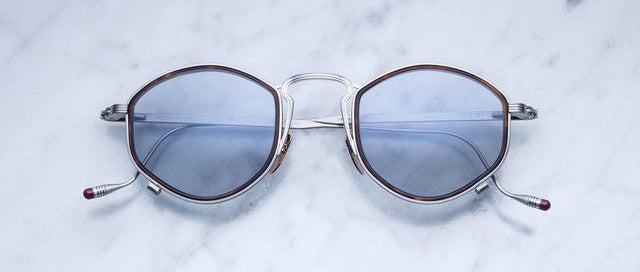A pair of round, silver-framed sunglasses with blue-tinted lenses rests on a white marble surface. The ends of the temples have small red tips.