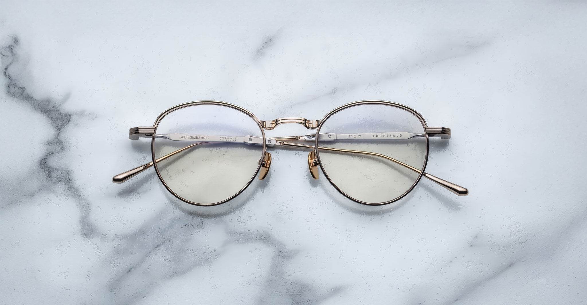 A pair of Jacques Marie Mage Archibald round, Altan light gold-framed optical frame with thin metal arms rests on a white marble surface with grey veining.