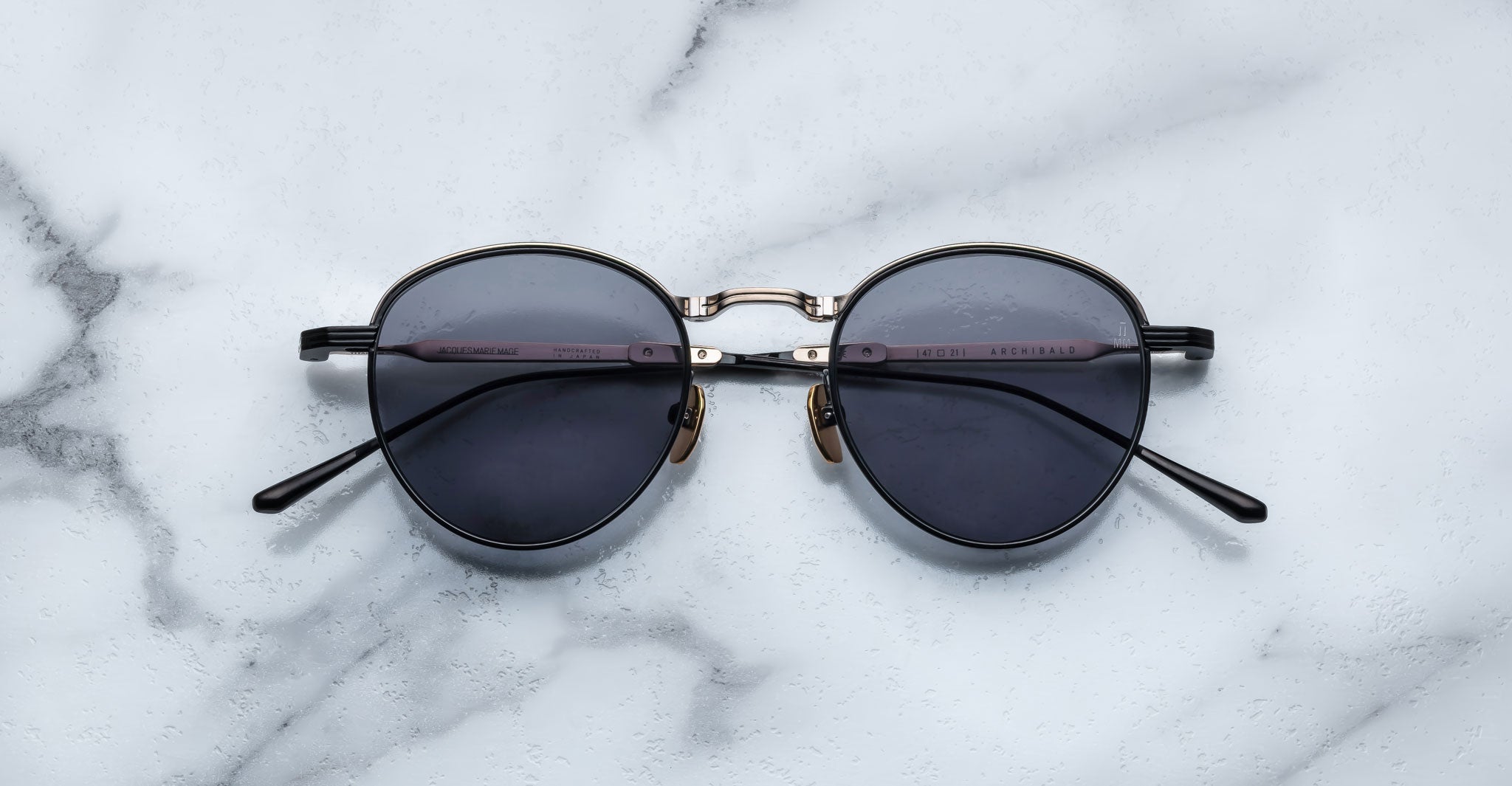 A pair of round, moon gold-framed sunglasses with charcoal lenses is placed on a white marble surface. The arms of the sunglasses are metallic with visible branding on the inner side of the arms.