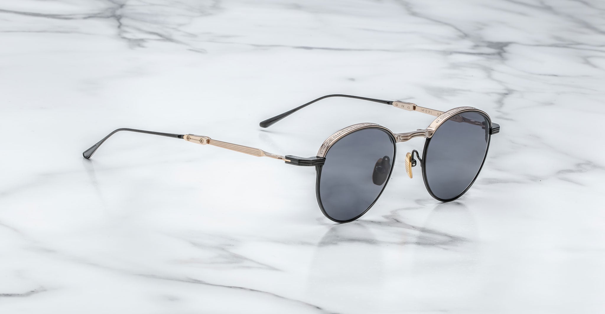 A pair of Jacques Marie Mage Archibald round, moon gold-framed sunglasses with thin metal arms rests on a white marble surface with grey veining.